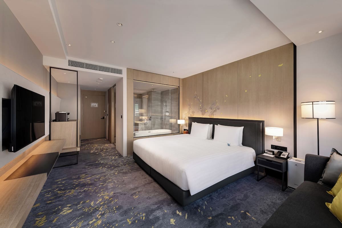 A modern hotel room with a large bed, a TV, a desk, and a bathroom visible through a glass partition.