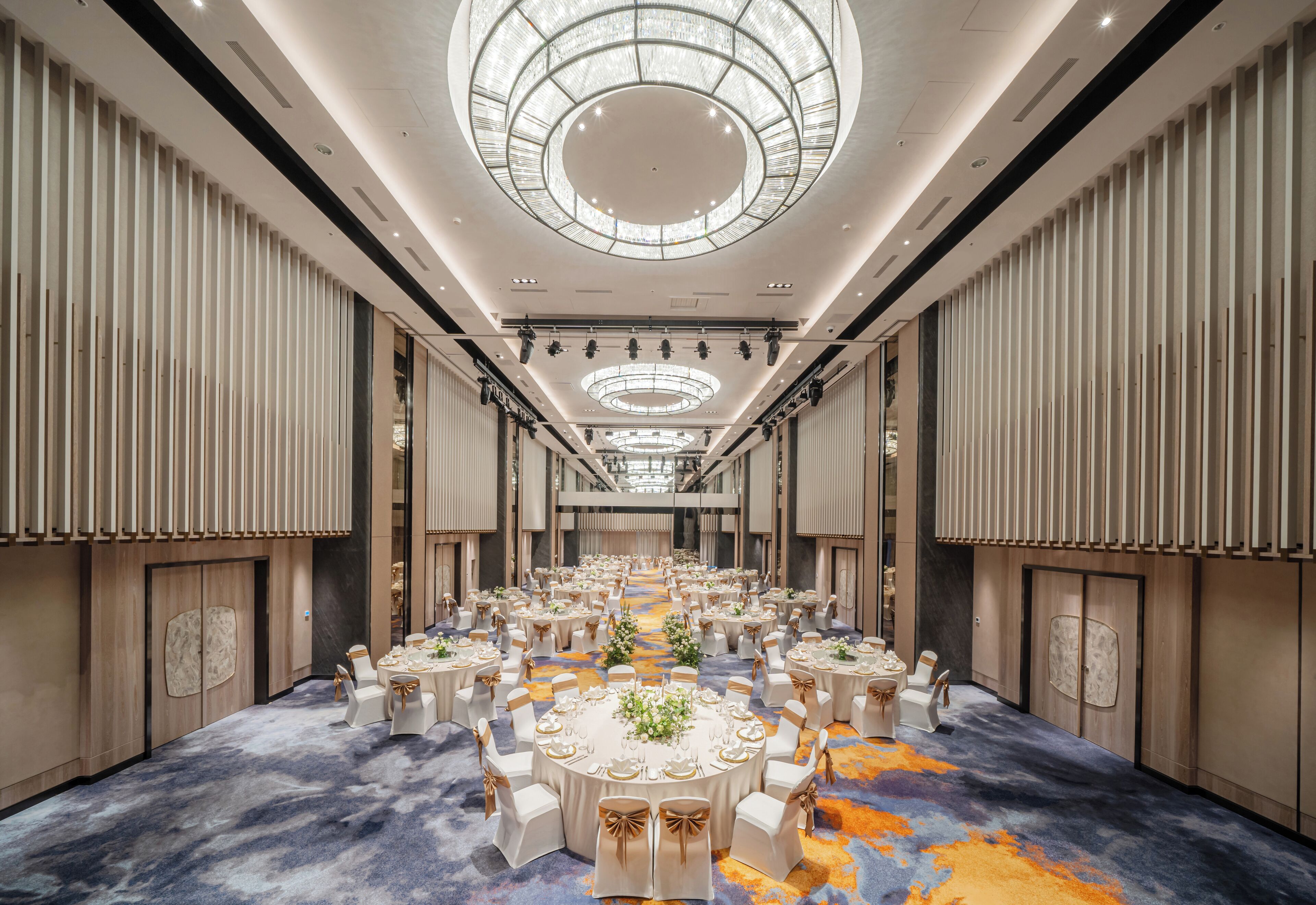 Une grande salle de banquet, bien éclairée, avec des tables et des chaises rondes, un arrangement floral central et un design de plafond moderne.