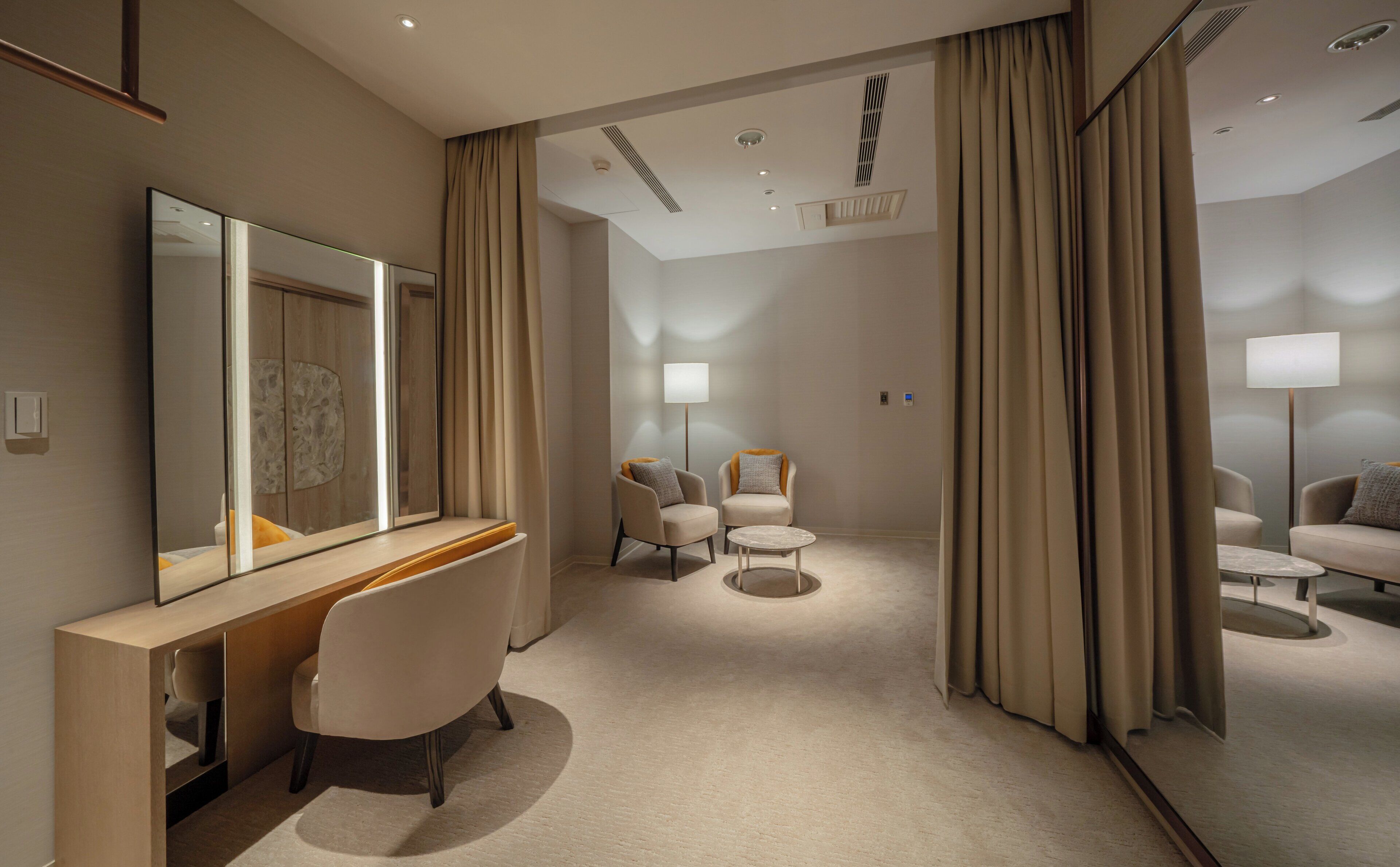 Habitación de hotel con un escritorio, dos sillones, una mesa redonda de centro, un espejo grande y una lámpara de pie.