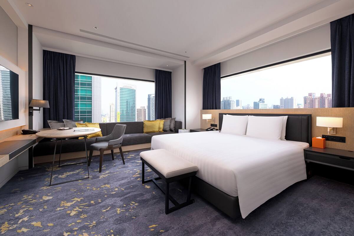 A hotel room with a bed, a desk, a chair, a bench, and a view of the cityscape.