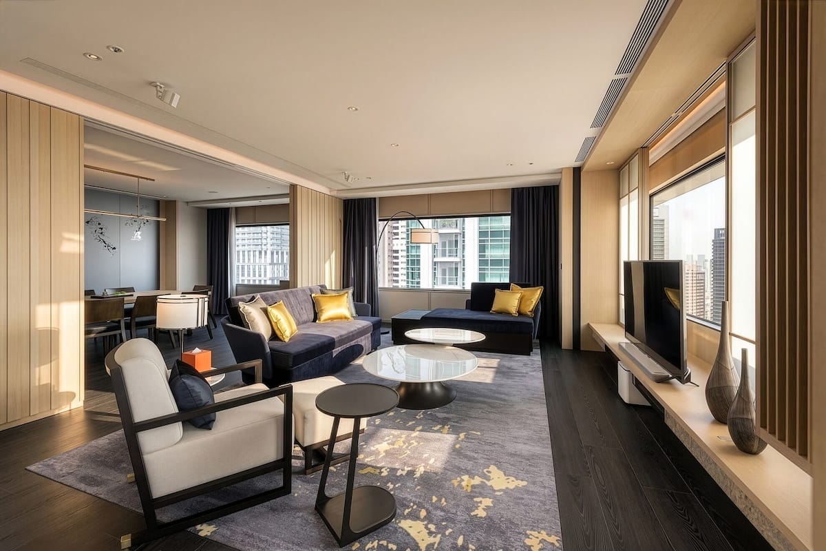 A modern hotel room with a sofa set, a dining area, a TV, and a large window with a city view.