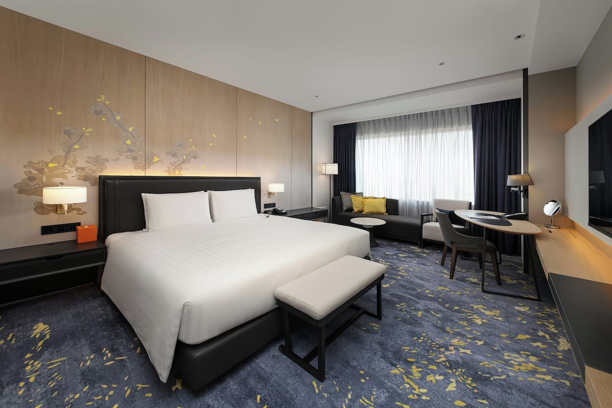 A hotel room with a large bed, bedside tables, a desk, a chair, a sofa, and a large window with curtains.