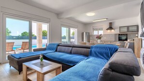 A modern living room with a large sectional sofa, a wooden coffee table, and a view of a swimming pool through large sliding glass doors. - Vacation home Kliska by Interhome (Pula)