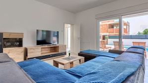 A modern living room with a large flat-screen TV, wooden furniture, and a view of a brick building through large windows. - Vacation home Kliska by Interhome (Pula)