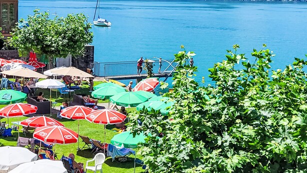 A lakeside terrace with outdoor seating and umbrellas, overlooking a calm lake and distant mountains.