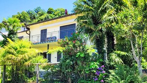 A two-story building with a balcony, surrounded by lush greenery and a stone wall.