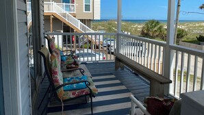A covered porch with a wooden deck, a white railing, and a view of the beach and ocean.
