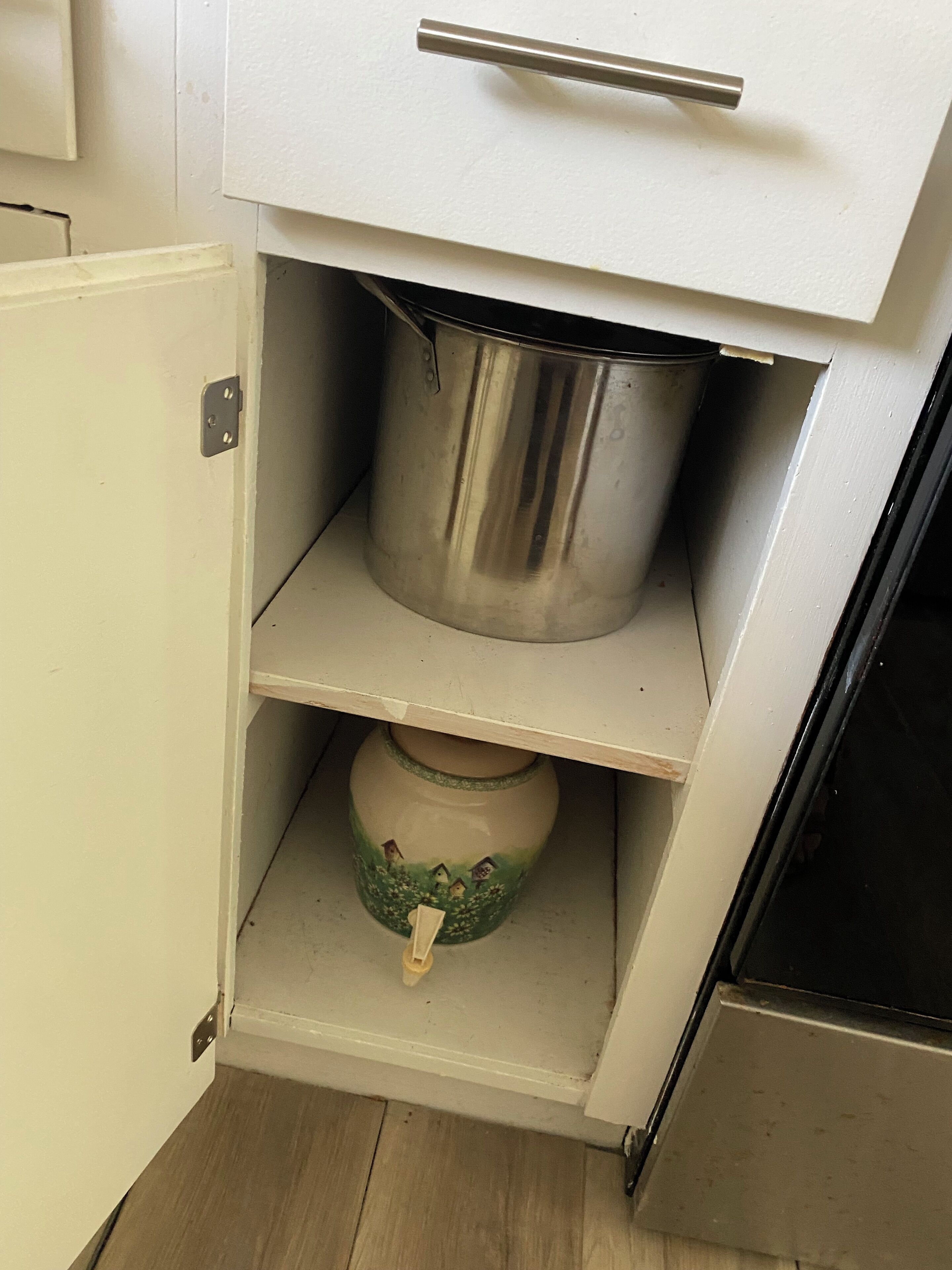 A kitchen with a white cabinet, a stainless steel pot on the shelf, and a ceramic pot with a floral design on the lower shelf.