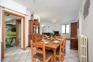 A dining area with a wooden table set for four, surrounded by chairs with cushioned seats. A cabinet with glass doors is visible, and a radiator is mounted on the wall.