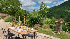 An outdoor dining area with a table set for breakfast, surrounded by lush greenery and a mountainous backdrop. - Olimpia by Interhome (Borgo San Lorenzo)