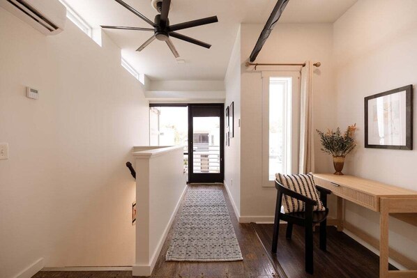 A hallway with a ceiling fan, a wooden floor, a rug, a chair, a table, and a door leading to another room.