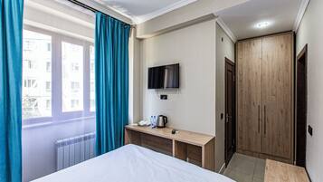 A hotel room with a bed, a desk with a flat-screen TV, a wooden wardrobe, and a window with curtains.