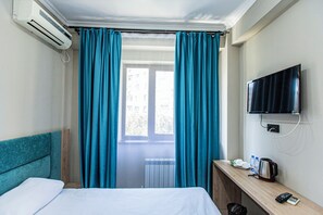 A hotel room with a bed, a desk with a kettle, a TV, and a window with curtains.