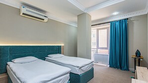 A hotel room with two beds, a ceiling air conditioner, a view of the outside, and a balcony with blue curtains.