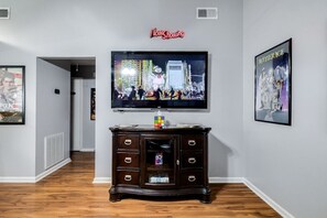 A living room with a flat-screen TV mounted on the wall, a dark wooden cabinet below it, and framed posters on the walls.