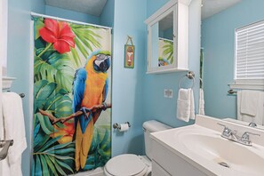 Un baño con un mural de guacamayo, un inodoro, un lavabo y una cortina de ducha con motivos tropicales.
