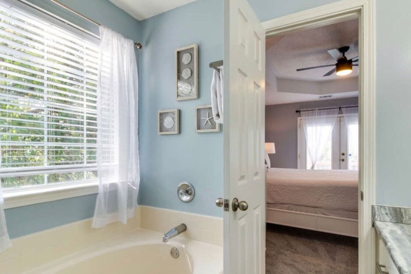 Un baño con una bañera, una ventana con persianas y una puerta que da a un dormitorio con una cama y un ventilador de techo.