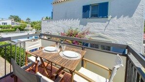 A balcony with a wooden table set for two, a white wall with blue shutters, and a clear blue sky.