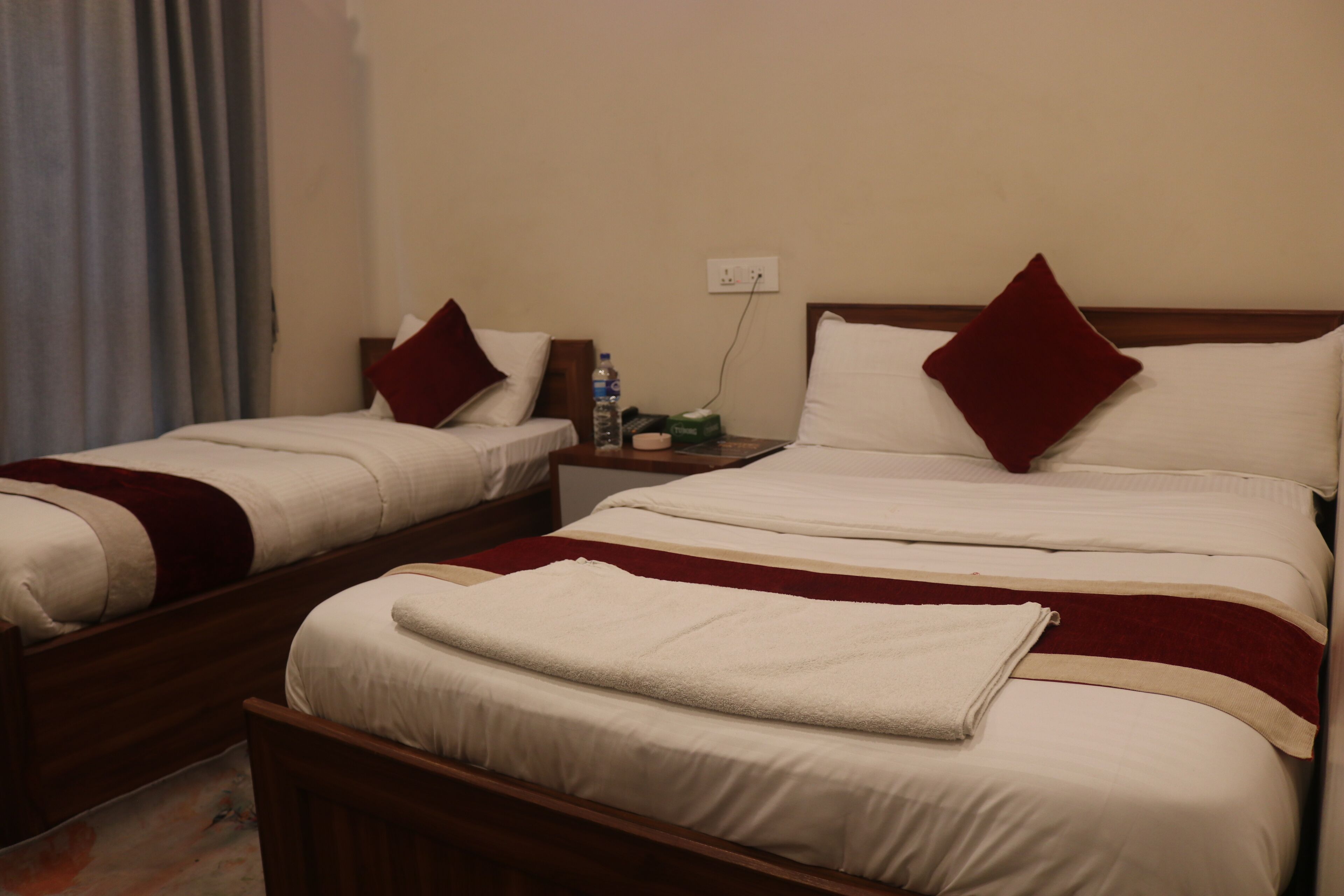 Two single beds with wooden frames, white bedding, and red pillows. There is a nightstand with a water bottle and a green box.