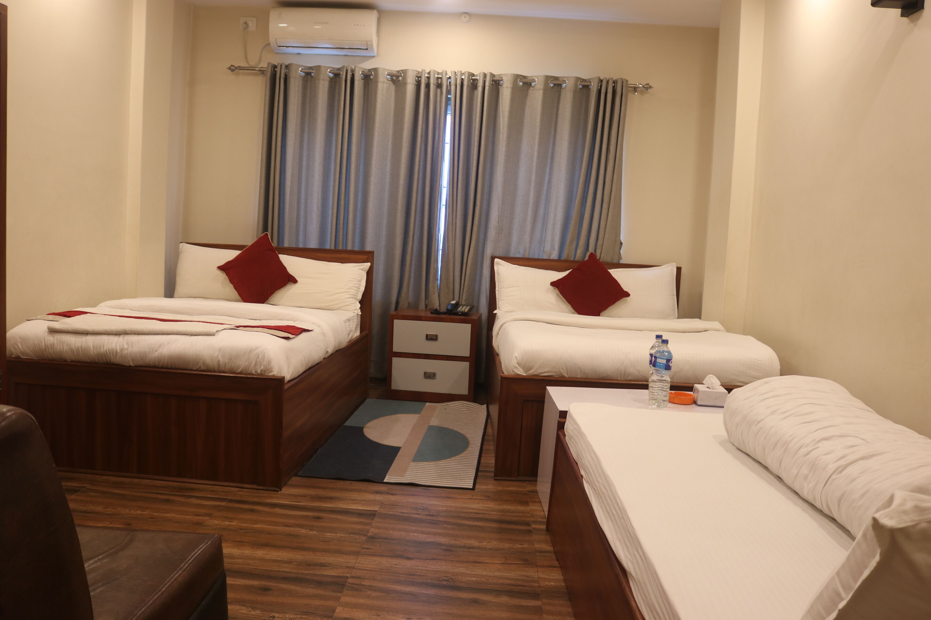 A hotel room with two beds, a wooden floor, a small bedside table with a lamp, and a window with curtains.