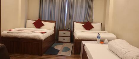 Habitación de hotel con dos camas, piso de madera, una mesita de noche pequeña con lámpara y una ventana con cortinas.
