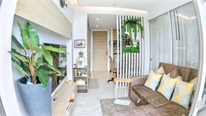 A modern living room with a sofa, a large potted plant, and a view into the kitchen area. - Rocco 513 - Corner 1 Bedroom Serviced Apartment With Seaview (Krabi)