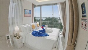 A hotel room with a large bed, a nightstand, a window with curtains, and a view of the outdoors. - Rocco 513 - Corner 1 Bedroom Serviced Apartment With Seaview (Krabi)