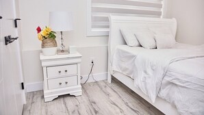 A white bedroom with a bed, a nightstand with a lamp, and a vase of flowers. - Cozy 3-bedroom, 3 queen beds ground level suite in a desirable location in PG.  (Prince George)