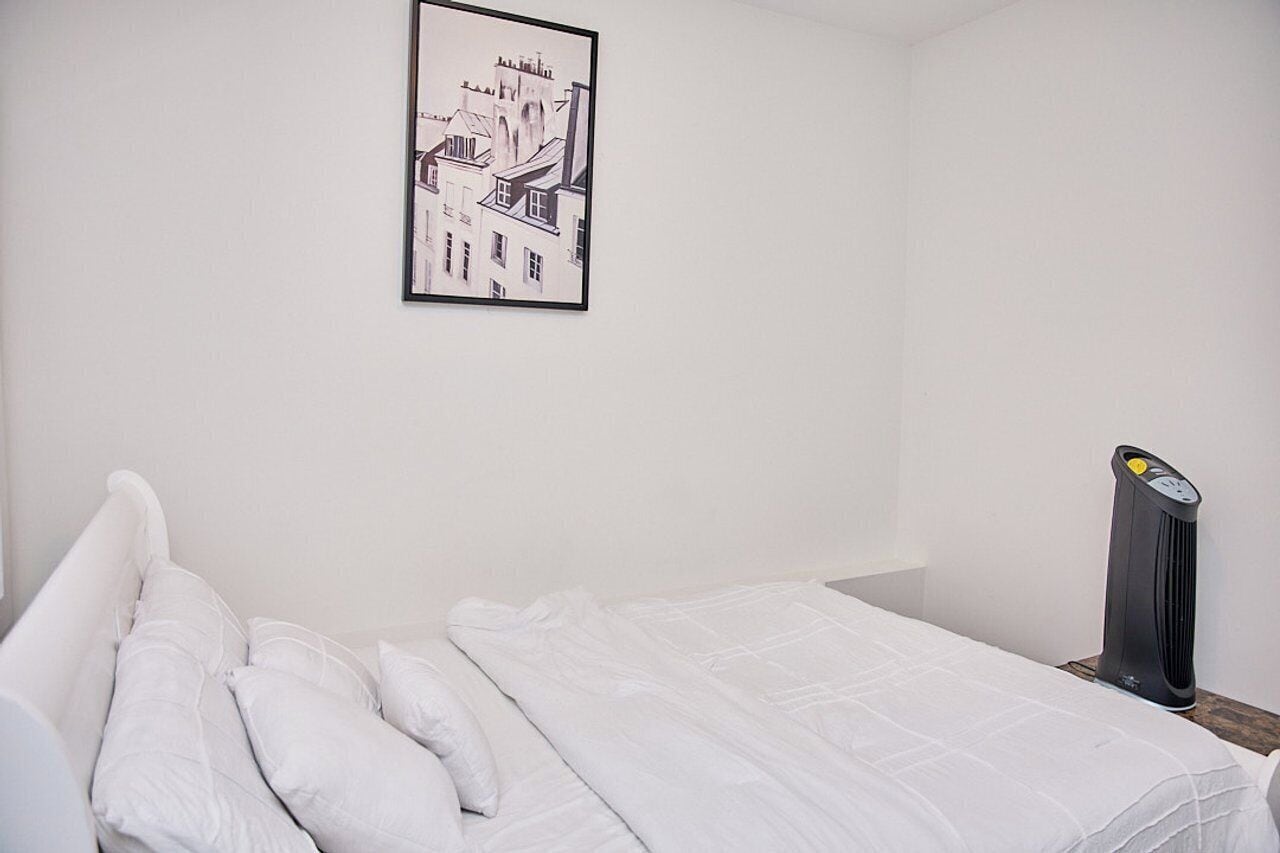 A neatly made bed with white bedding, a framed black and white cityscape picture on the wall, and a black heater on the nightstand.