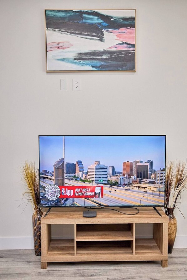 Un televisor sobre un soporte de madera, con un fondo de paisaje urbano, un cuadro abstracto enmarcado encima y dos jarrones decorativos a cada lado.