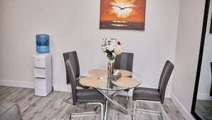 A dining area with a glass table, four chairs, a vase with flowers, a water dispenser, and a painting on the wall. - Cozy 3-bedroom, 3 queen beds ground level suite in a desirable location in PG.  (Prince George)