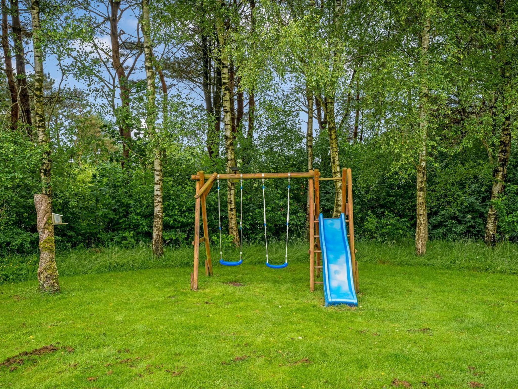 A wooden swing set with two swings and a blue slide in a grassy area surrounded by trees.
