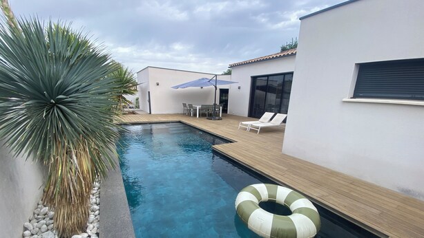 Un espace piscine extérieur moderne, doté d’une terrasse en bois, de chaises longues et d’un palmier.