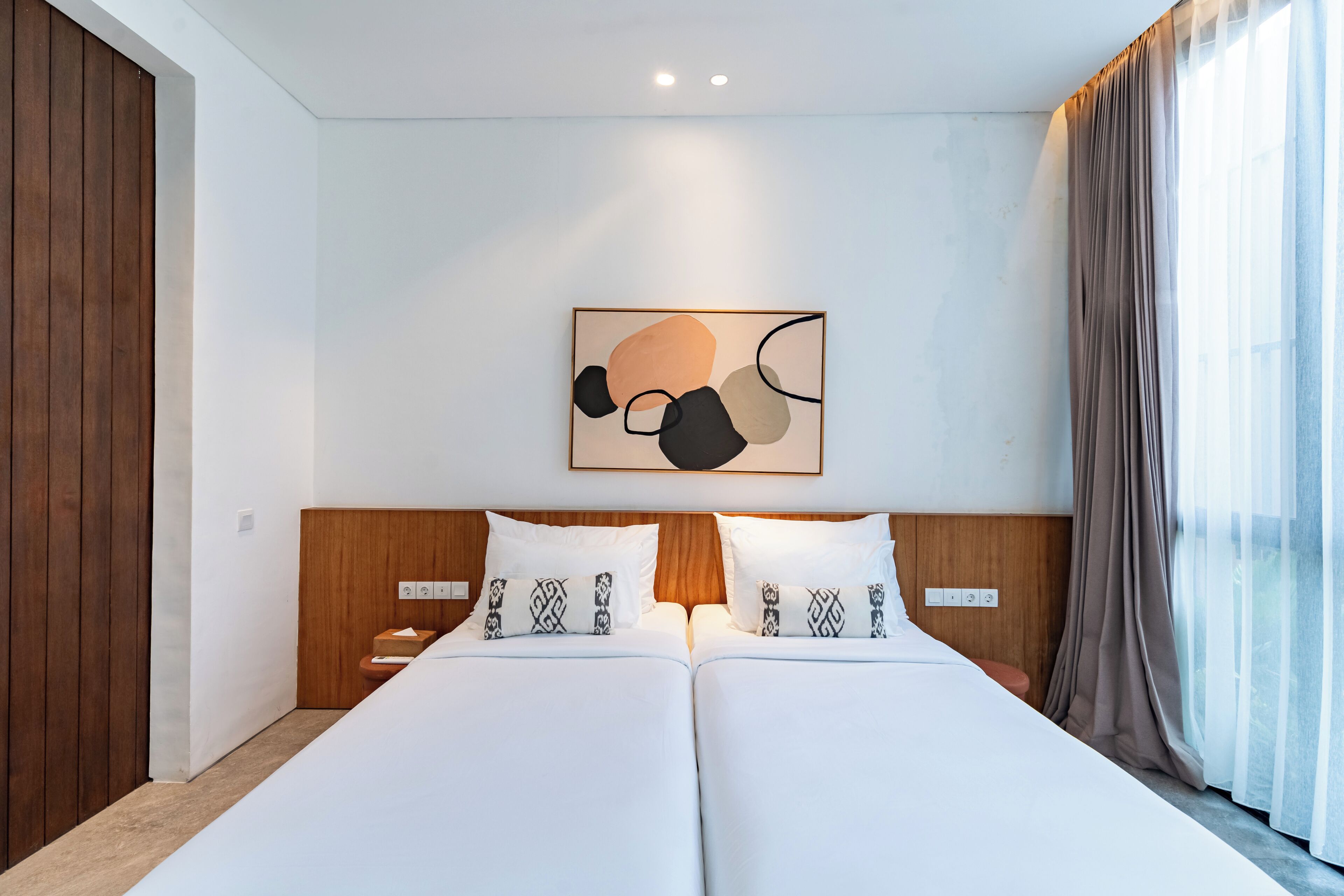 A double bed with white linens and patterned pillows, a wooden headboard, and a wall-mounted abstract art piece.