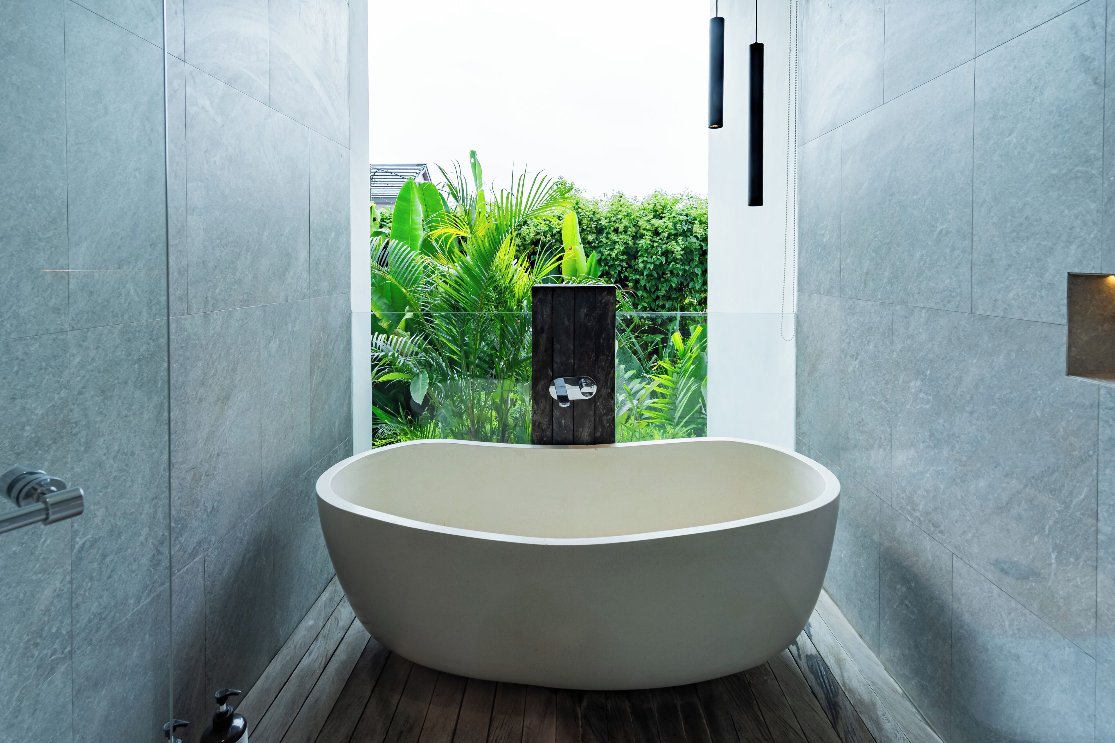 A modern bathroom with a freestanding bathtub, a wooden post, and a view of greenery outside.