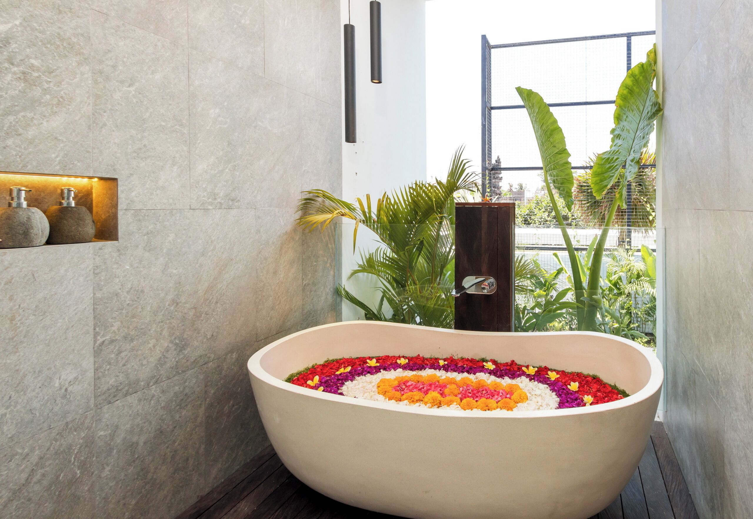 A large bathtub filled with colorful flowers, a wooden shelf with bottles, and a view of greenery outside.