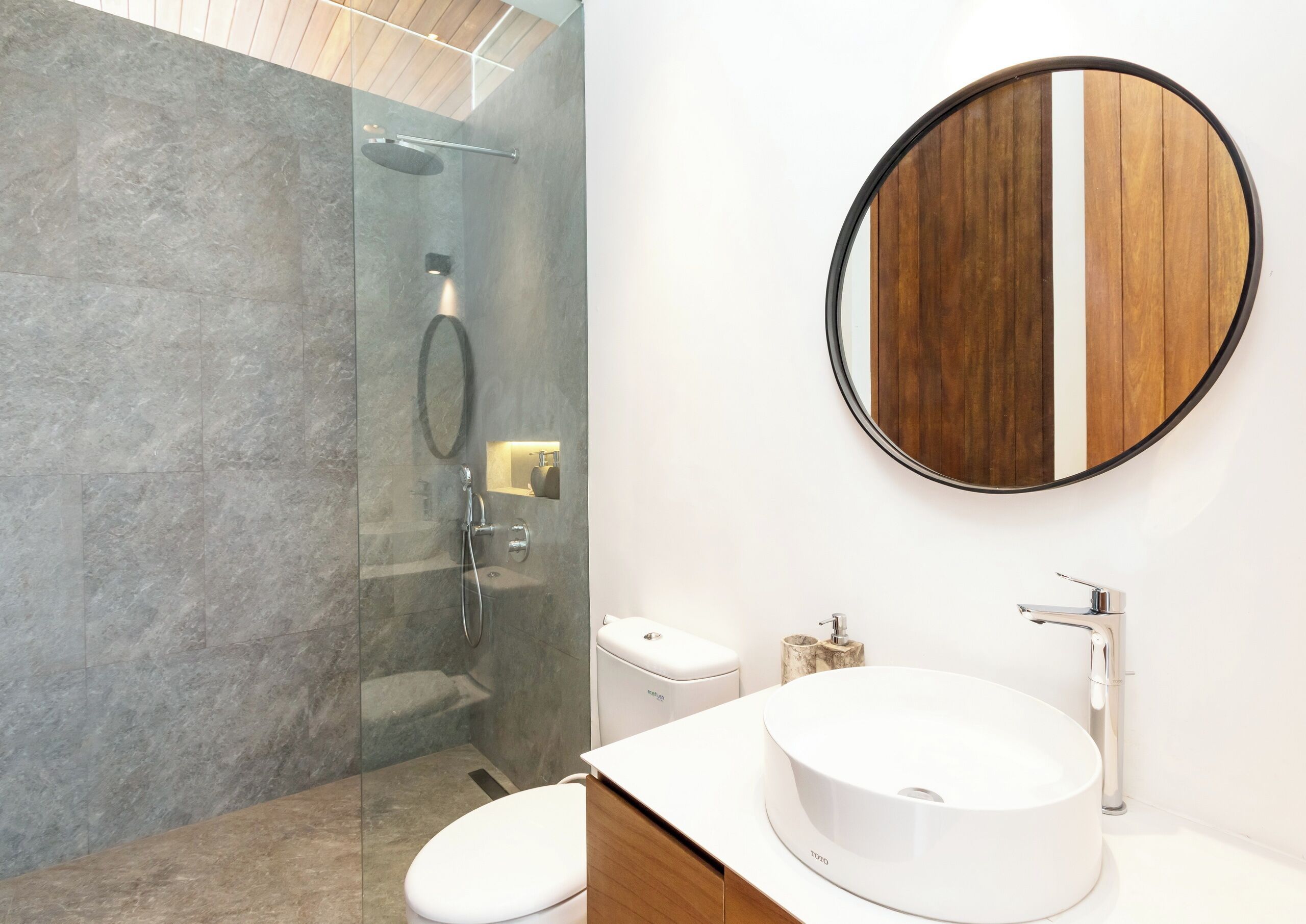 A modern bathroom with a glass-enclosed shower, a round mirror, a white sink, and a toilet.