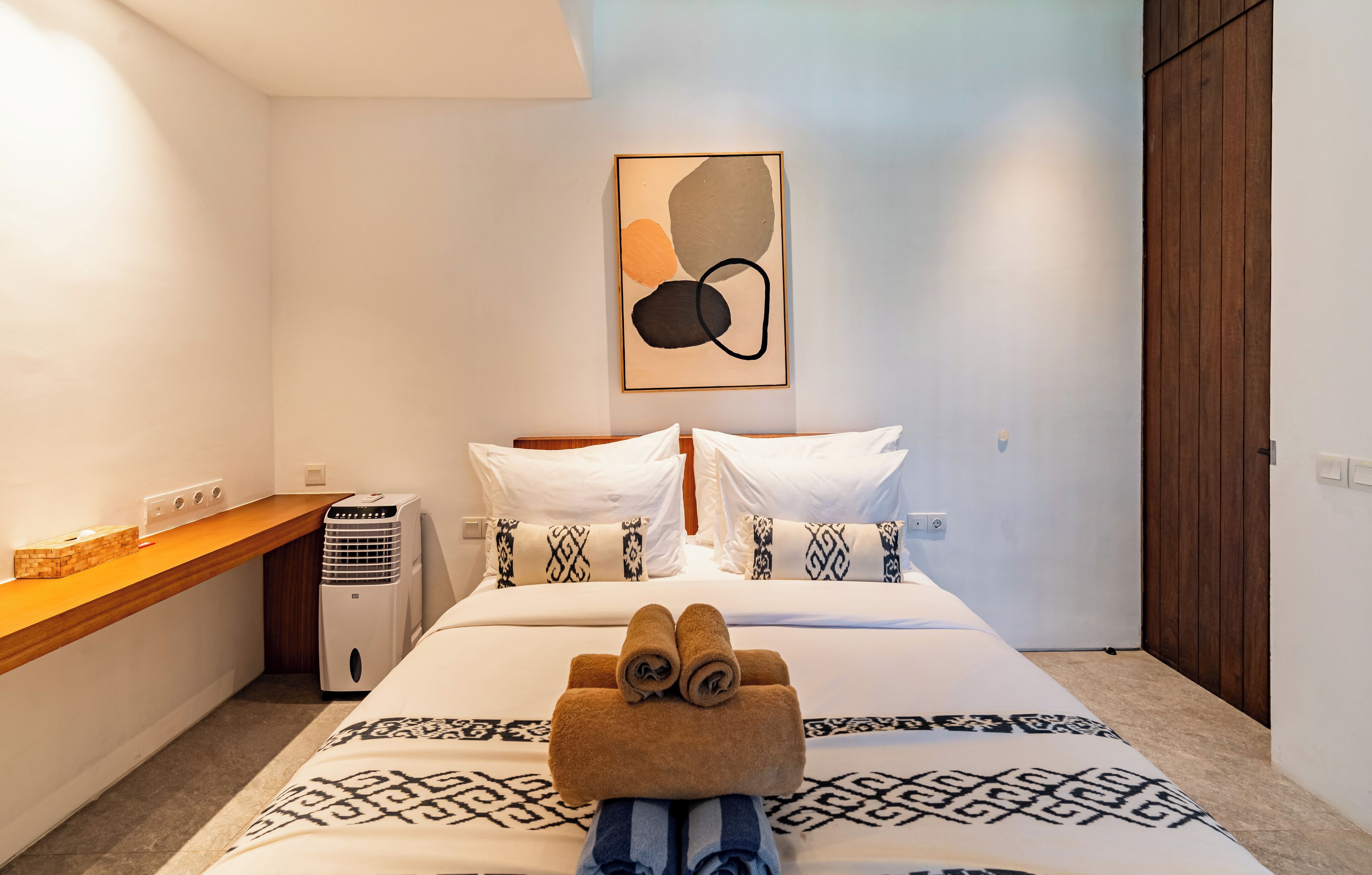 A hotel room with a neatly made bed, a wooden bedside table, a wall-mounted air conditioner, and a framed abstract artwork above the bed.