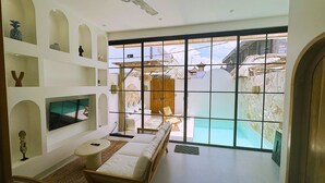 A modern room with a large window, a ceiling fan, a TV, and a seating area with a view of a pool and outdoor structures. - Playa Canggu 7 (Canggu)