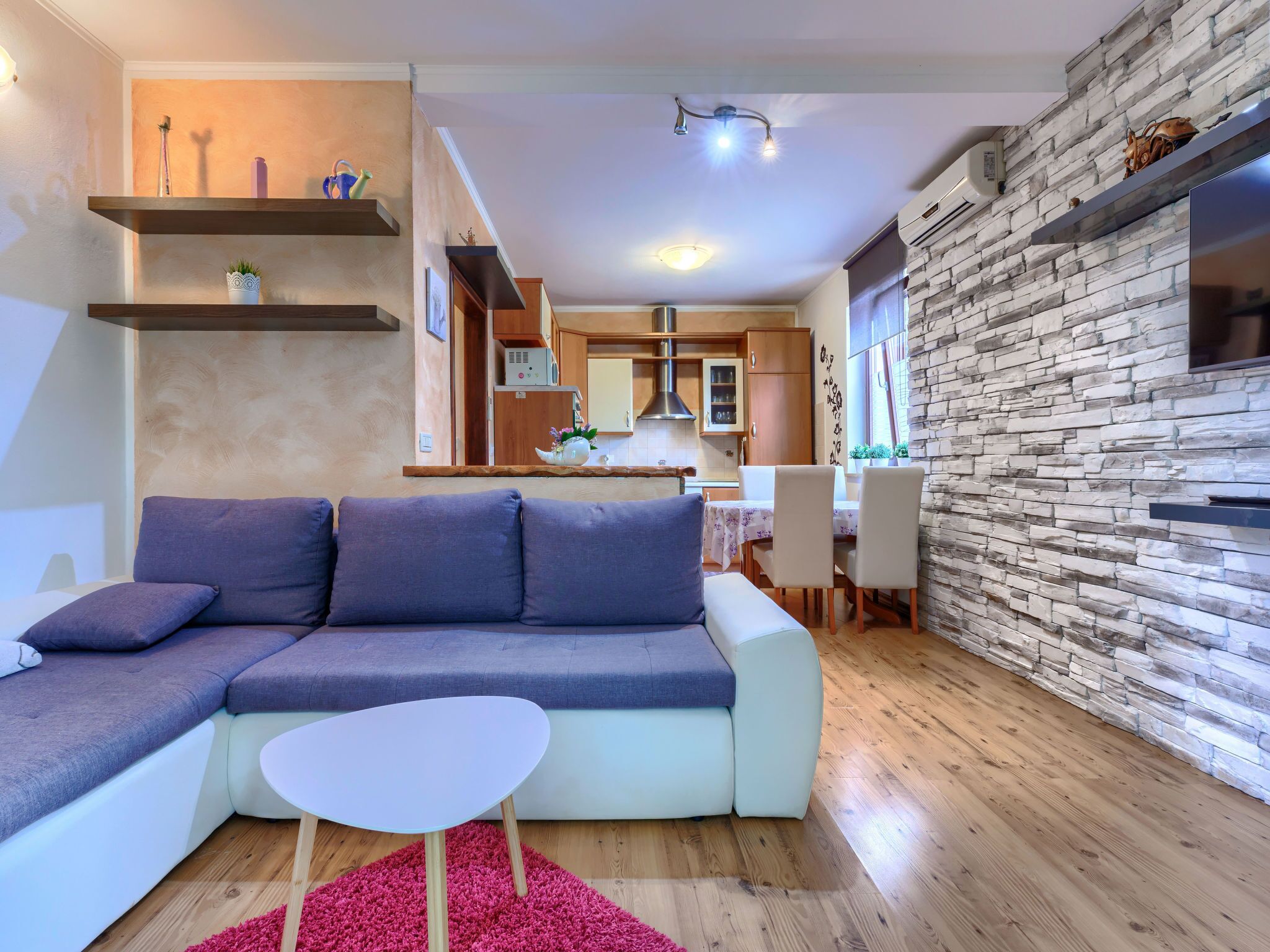 Ein modernes Wohnzimmer mit einem Ecksofa, einem kleinen runden Tisch und einer Küchenzeile mit Wandverkleidung aus Stein und Holzschränken.