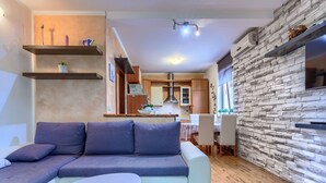 Un salon moderne avec un canapé d’angle, une petite table ronde et un coin cuisine doté d’un mur recouvert de pierres et de meubles en bois.