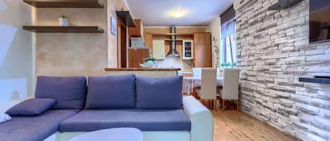 Ein modernes Wohnzimmer mit einem Ecksofa, einem kleinen runden Tisch und einer Küchenzeile mit Wandverkleidung aus Stein und Holzschränken.