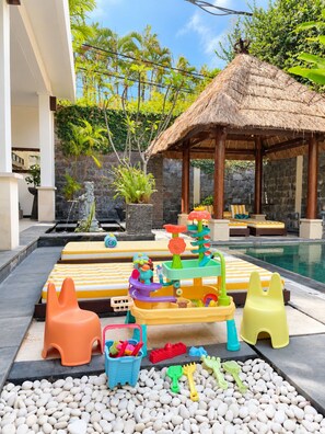 Zona de piscina con una estructura de techo de paja, juegos infantiles de colores y una estatua.