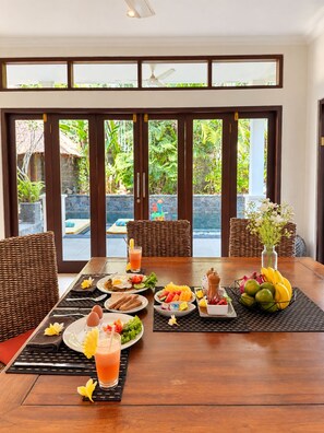Una mesa de comedor lista con alimentos para el desayuno, vista a un jardín exuberante a través de amplias ventanas y mobiliario de mimbre.