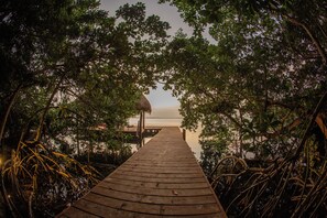 Een houten pad dat naar een strand leidt, omgeven door dichte begroeiing en een constructie met rieten dak.