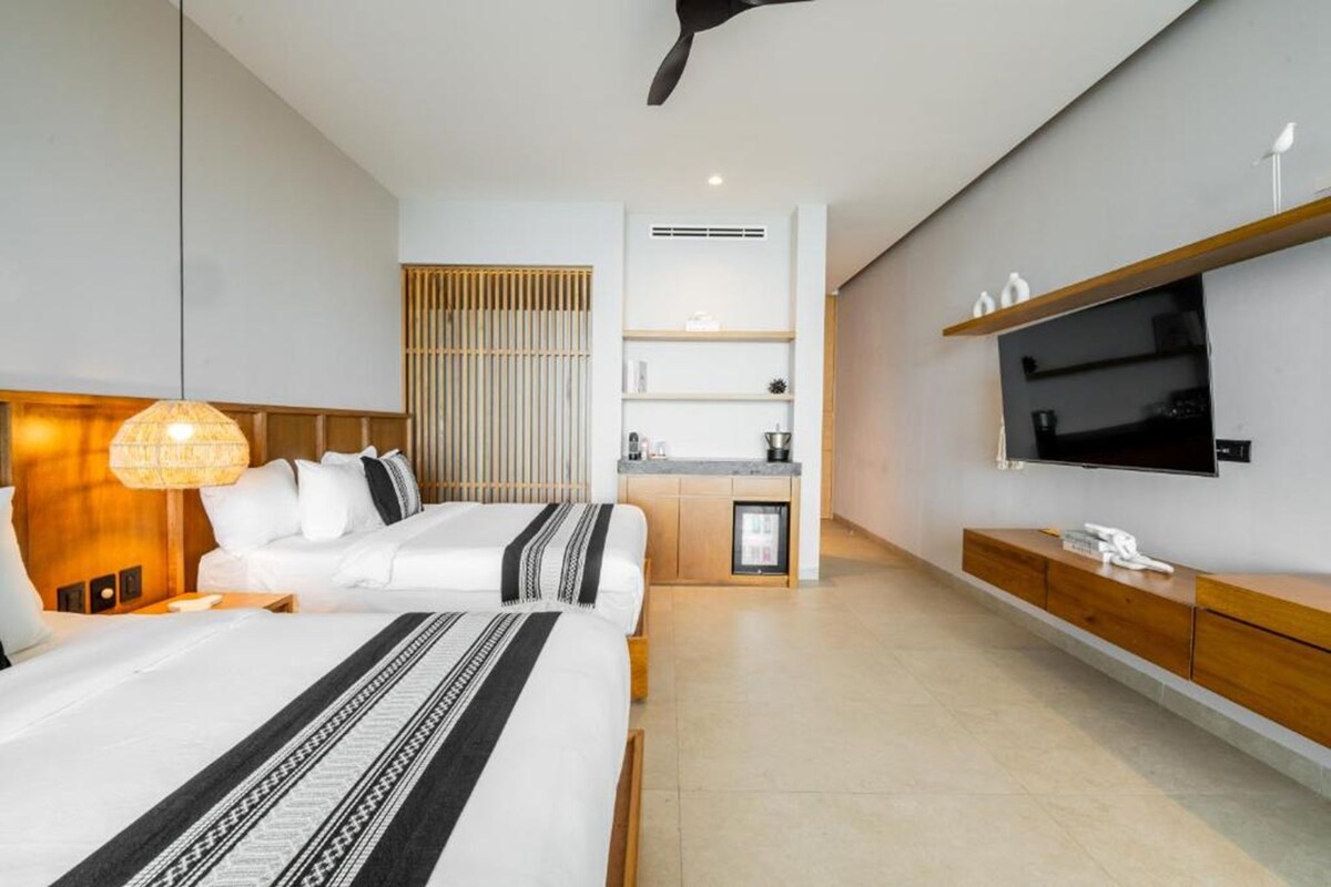 A hotel room with two beds, a kitchenette, a TV mounted on the wall, and a ceiling fan.