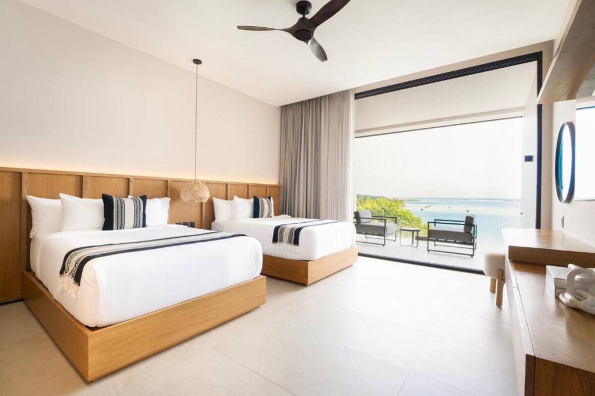 A hotel room with two beds, a desk, a chair, a ceiling fan, and a view of the ocean.