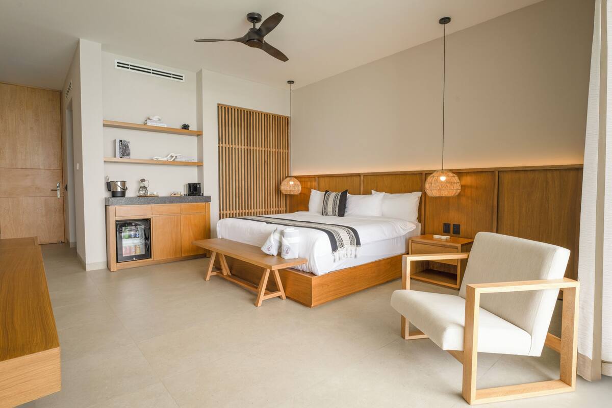 A modern bedroom with a wooden bed, bedside tables, a chair, and a built-in kitchen area.
