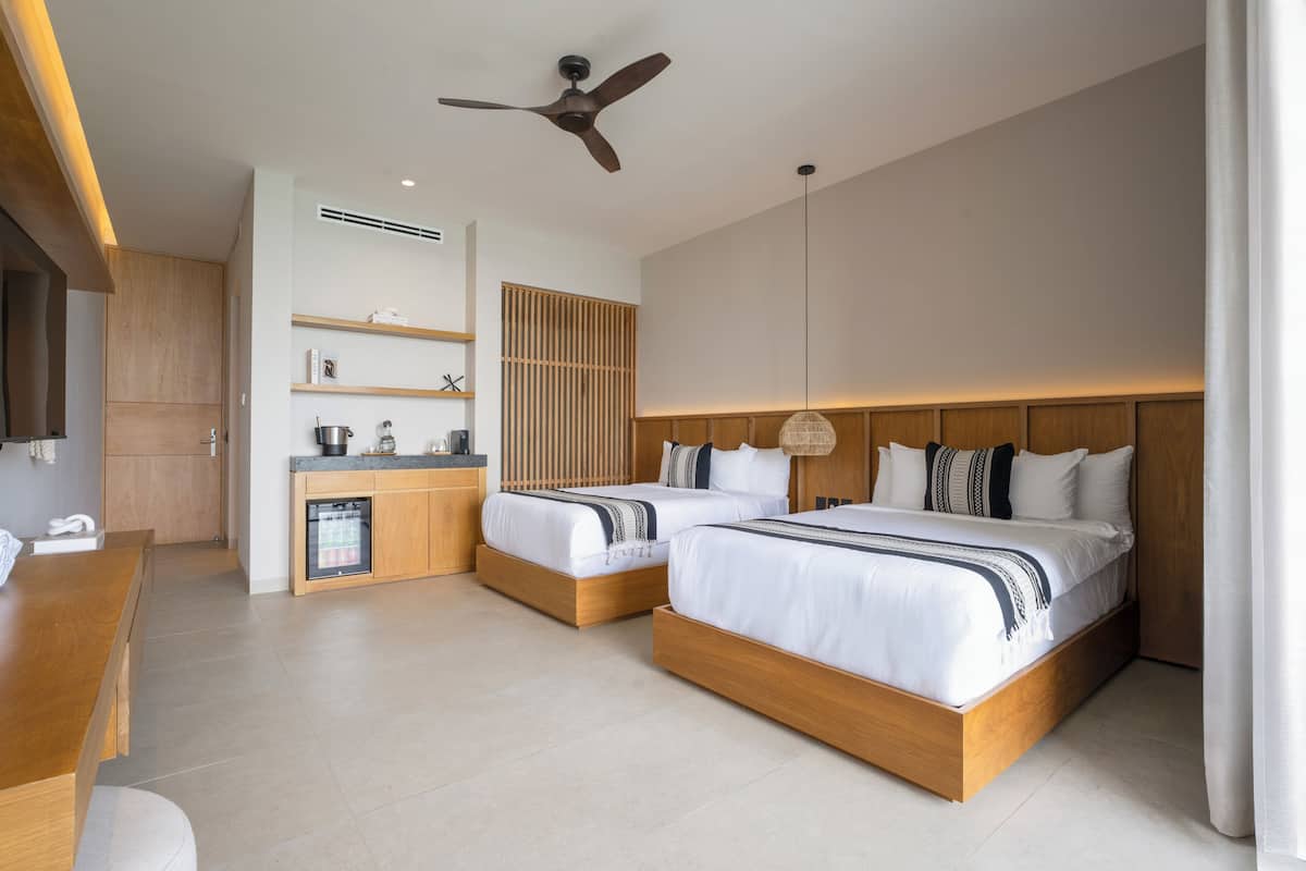 A modern hotel room with two beds, a wooden desk, a built-in kitchen, and a ceiling fan.