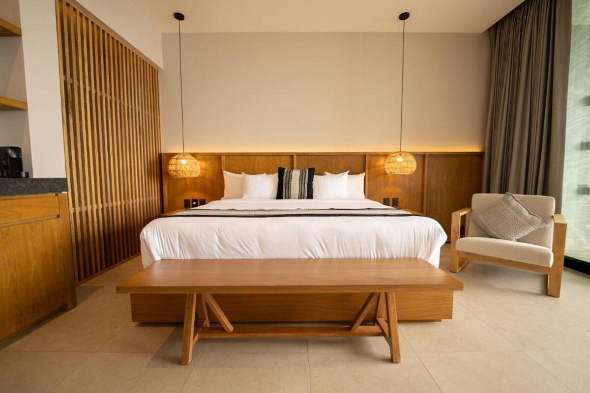 A modern hotel room with a large bed, bedside lamps, a wooden bench, a chair, and a view of the outdoors.